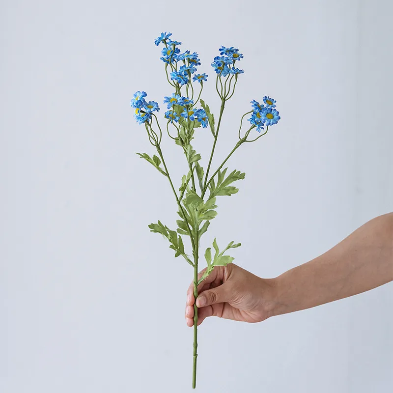 Blue artificial daisy spray stem with open blooms, buds and feathery foliage, held in one hand