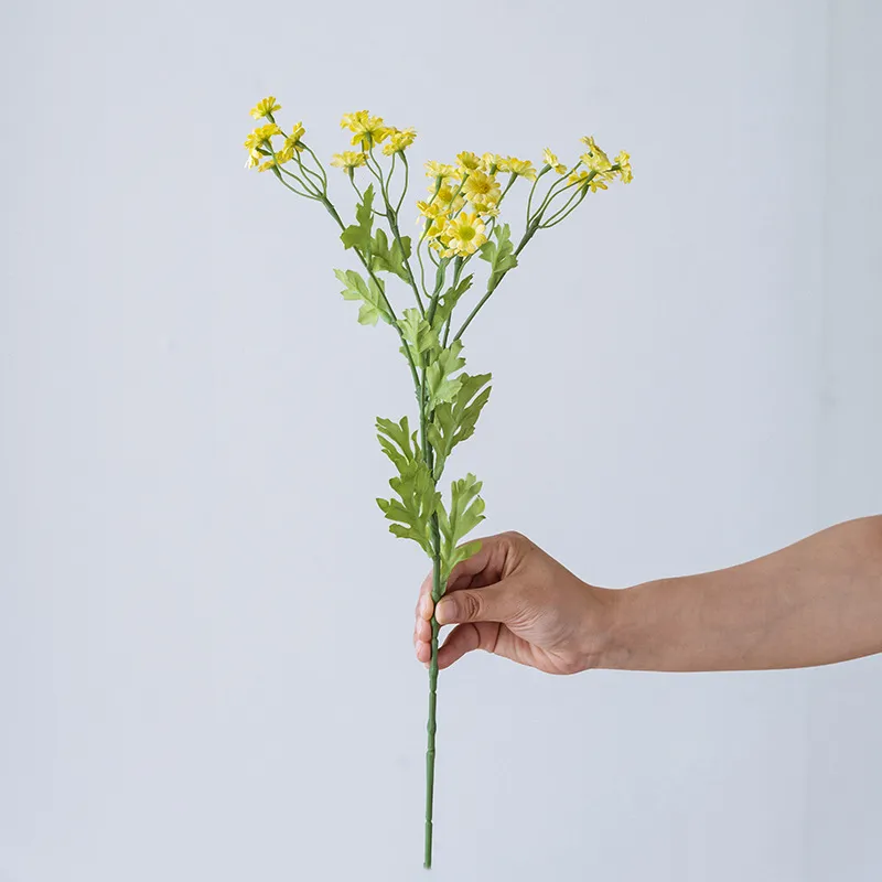 Yellow artificial daisy spray stem with open blooms, buds and feathery foliage, held in one hand