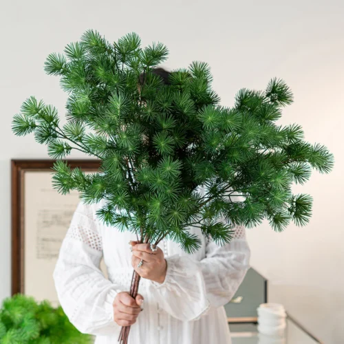 Bright green artificial pine branch with soft needled side shoots on a brown stem, held in one hand