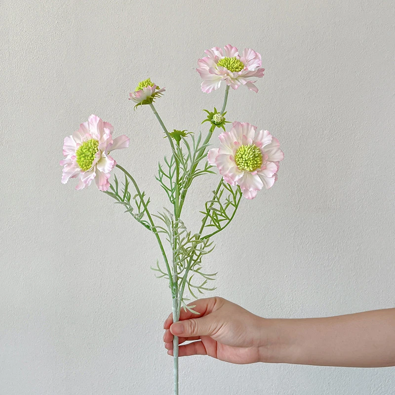 Blush artificial scabiosa stem with a lime-green pincushion centre and feathered leaves, held in one hand