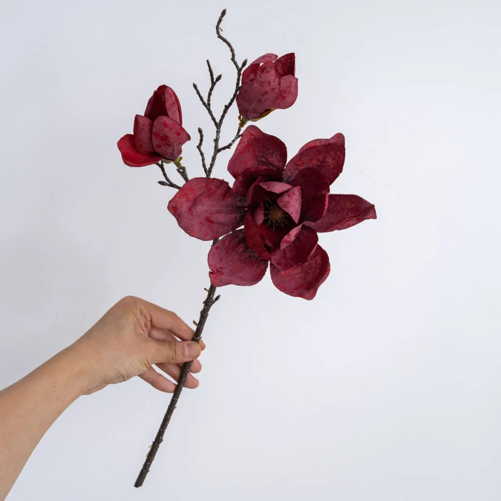 Burgundy dried look artificial magnolia stem on a bare twiggy branch, held in one hand