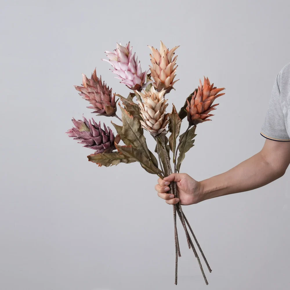 Dried look artificial protea stem with layered bracts and long leaves, held in one hand