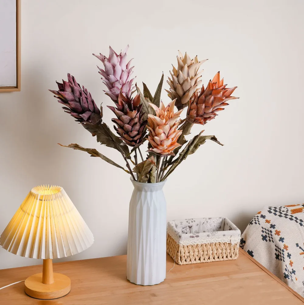 Dried look artificial protea stem with layered bracts and long leaves, styled in a room setting