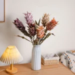 Dried look artificial protea stem with layered bracts and long leaves, styled in a room setting