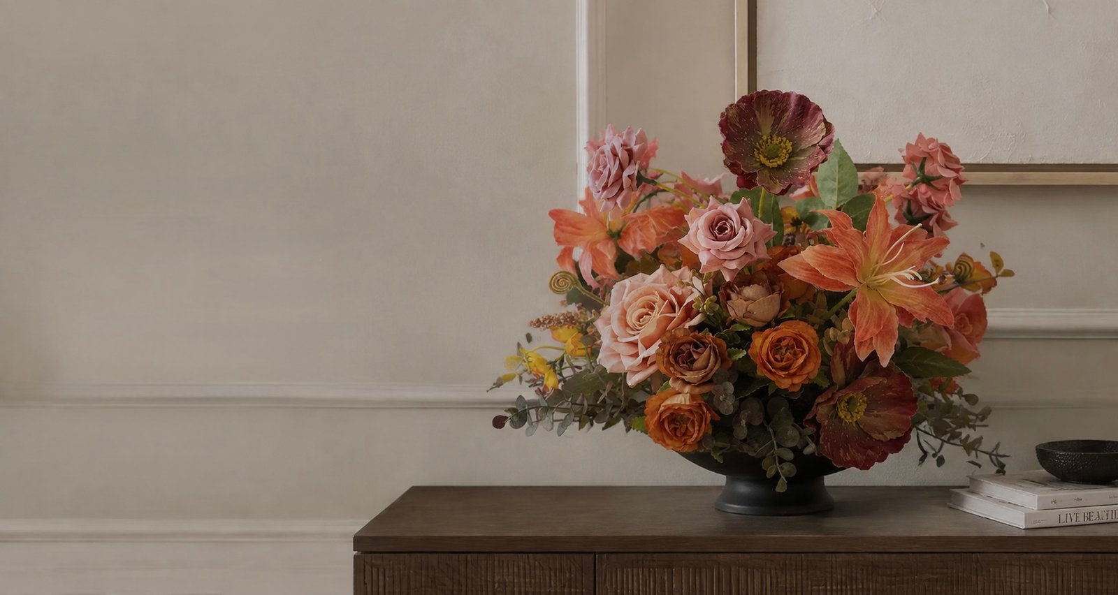 Dusty rose and terracotta faux flower arrangement in a black bowl on a dark wood credenza
