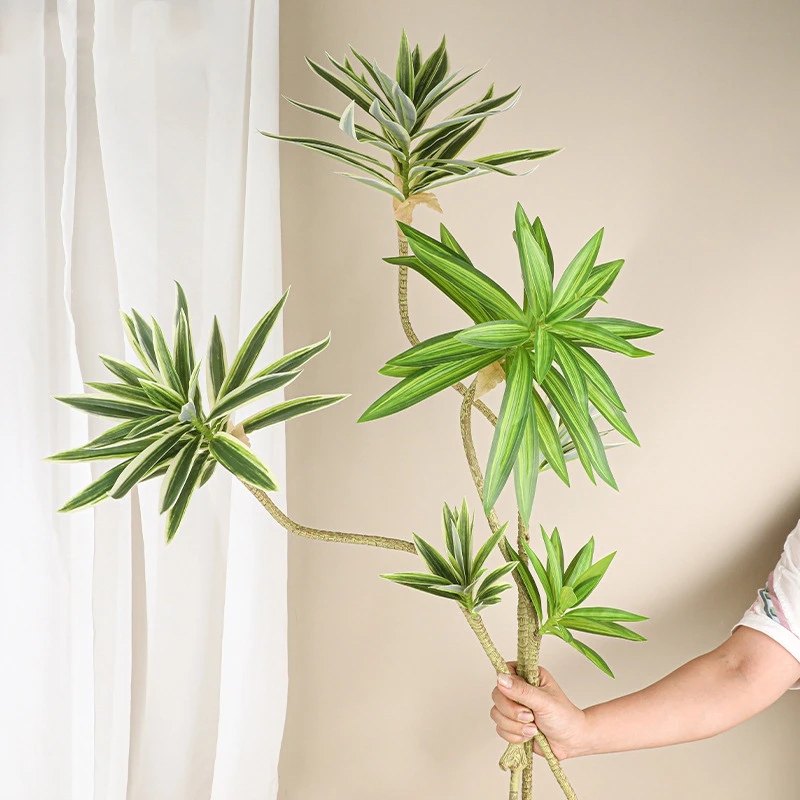 Tall, held artificial cordyline stem with narrow strap leaves on a curving bare stem, held in one hand