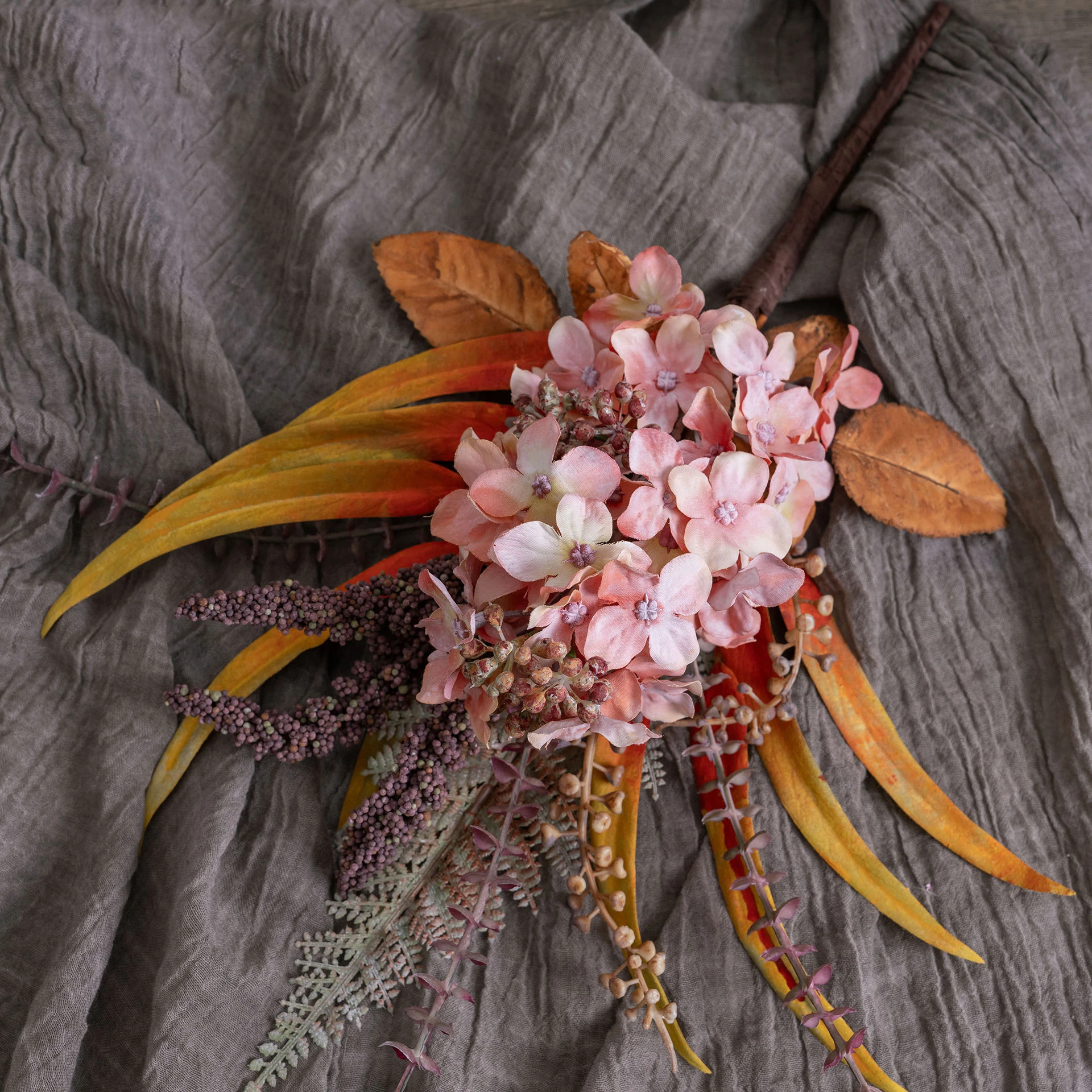Coral and rust artificial hydrangea pick built round one hydrangea head with autumn foliage behind it, styled on a table top