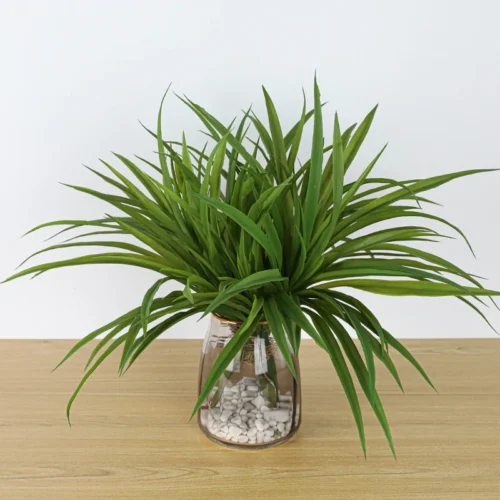 In a glass pot artificial tall grass plant as a dense tuft of long narrow blades, styled in a room setting