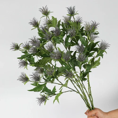 A full bunch artificial thistle flower stem with soft lilac crowns sitting above jagged leaves, held in one hand