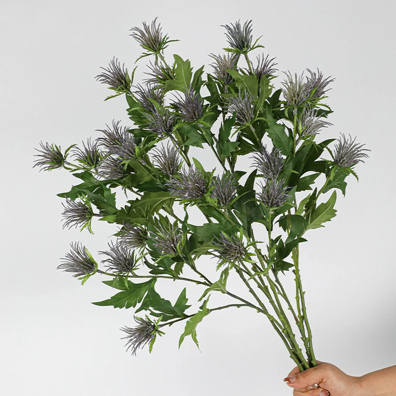 A full bunch artificial thistle flower stem with soft lilac crowns sitting above jagged leaves, held in one hand
